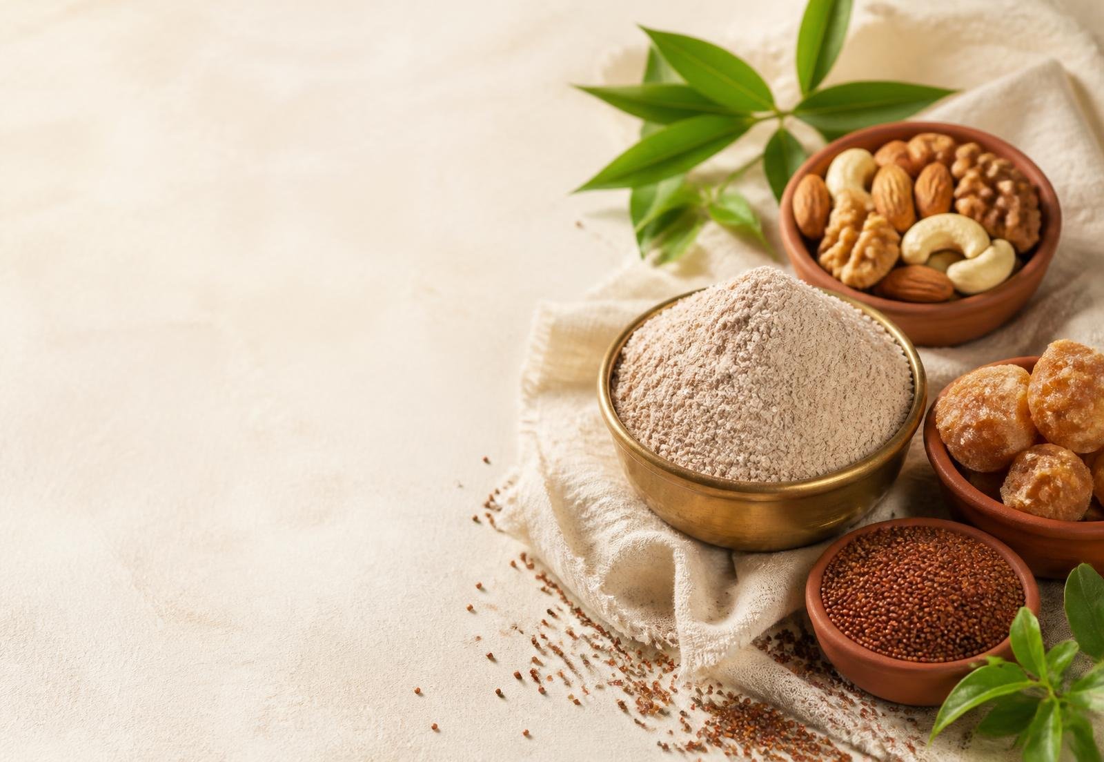 Brass bowl of finger millet flour with terracotta bowls of jaggery, dry fruits and ragi grains on cream linen